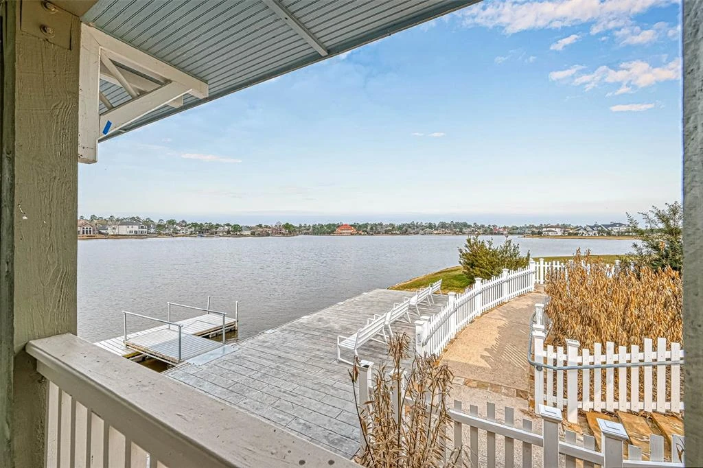 View from a covered lakeside structure toward a deck, white fencing, and homes across the water.