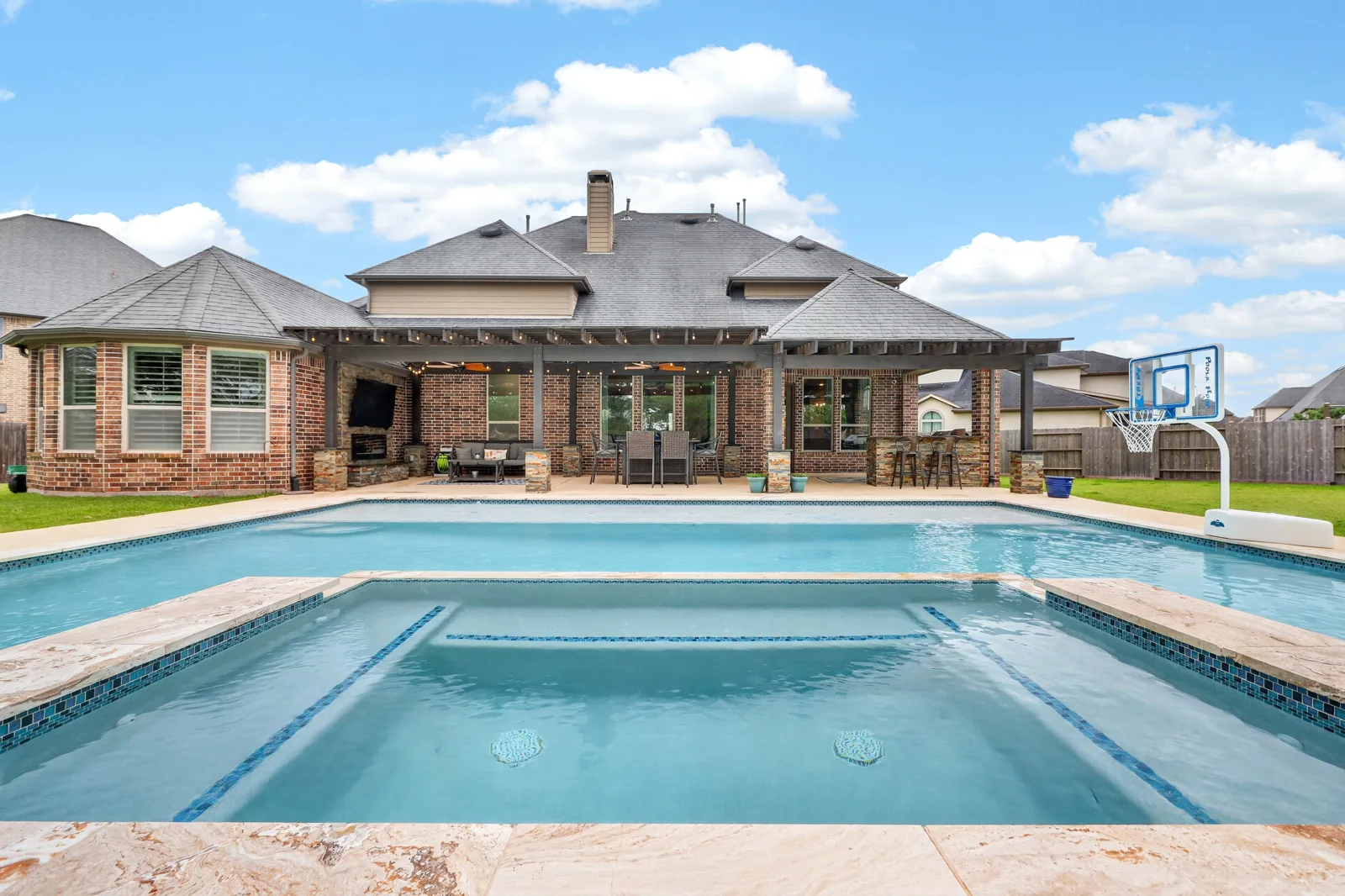 Backyard pool and spa in front of a brick home with a covered patio and outdoor seating.